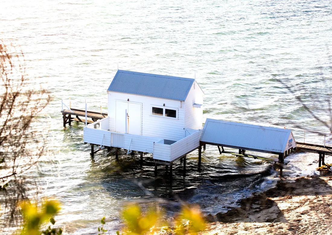 Small white boatshed on stilts by a body of water with a dock.