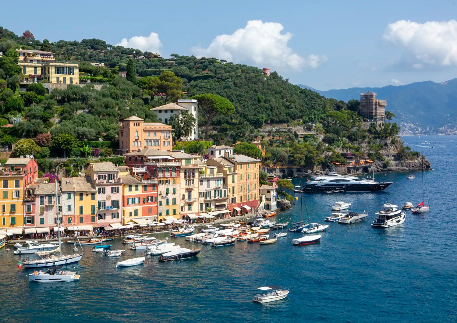 Coastal town with colorful buildings and boats in a harbor, surrounded by green hills and blue sky.