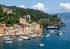 Coastal town with colorful buildings and boats in a harbor, surrounded by green hills and blue sky.