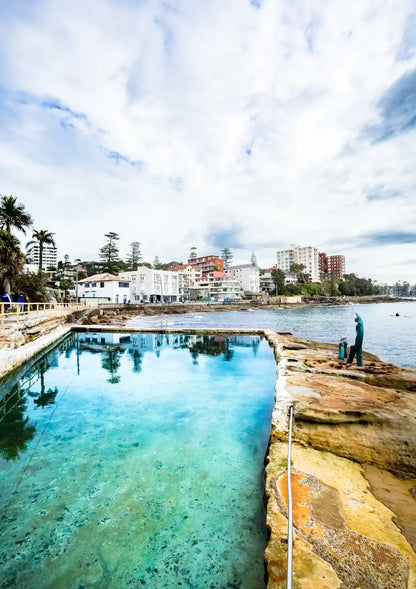 Coastal area with a pool of water, rocky shoreline, and buildings in the background.