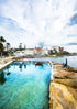 Coastal area with a pool of water, rocky shoreline, and buildings in the background.