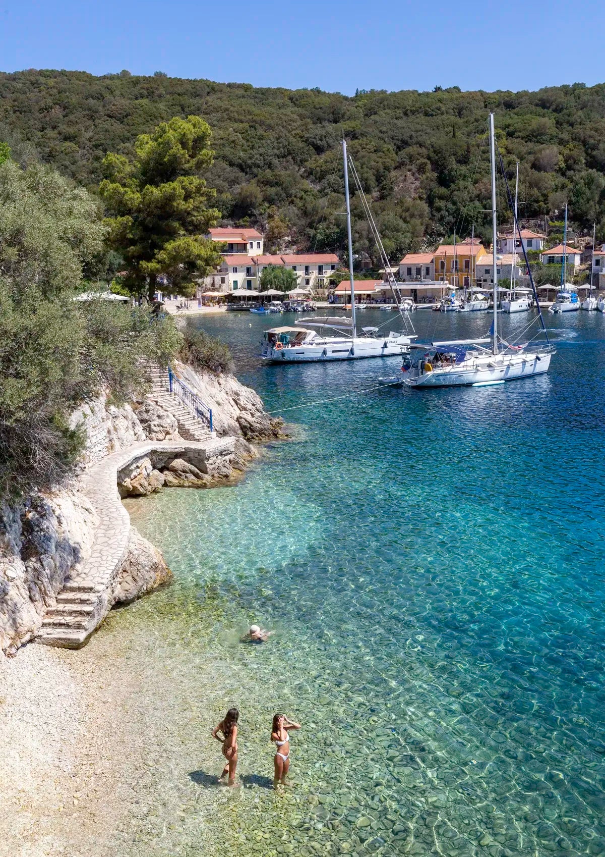 Two girls in bathers standing in clear water near a rocky shore with boats and buildings in the background.