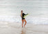 Person in a black swimsuit holding a green surfboard on a beach with waves.