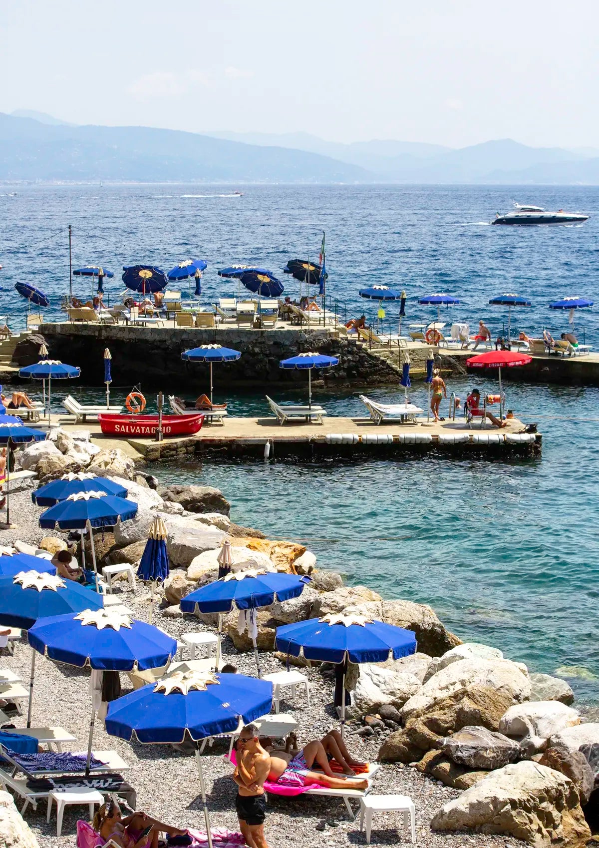 Beach scene with blue umbrellas, lounge chairs, and people near a dock with boats.