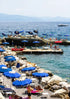 Beach scene with blue umbrellas, lounge chairs, and people near a dock with boats.