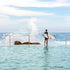 Person holding a surfboard standing in shallow water with a large wave crashing behind them.