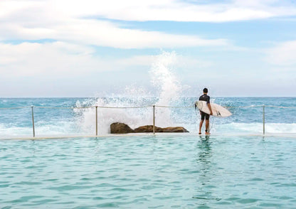 Person with a surfboard standing in a pool of water looking at the ocean.