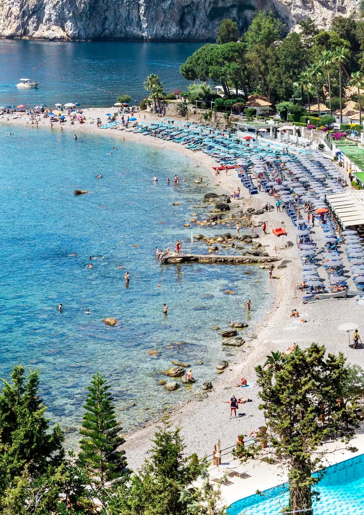Beach scene with people, umbrellas, and boats near a rocky coastline.