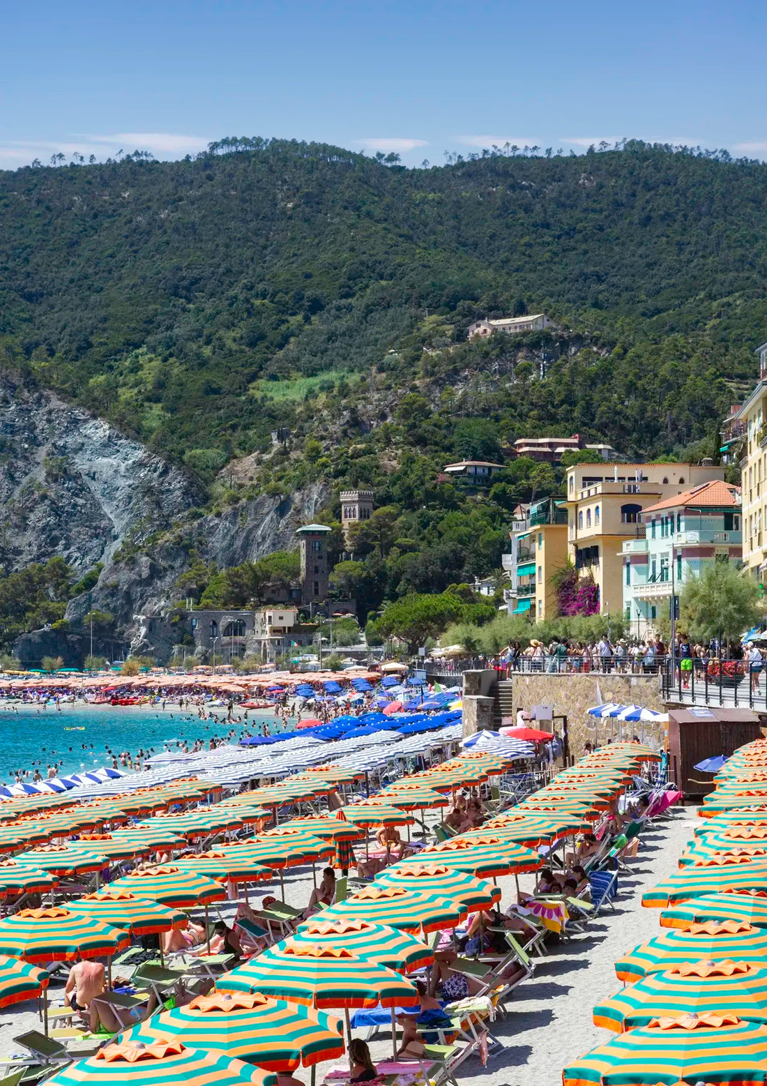 Beach scene with rows of striped umbrellas and people, surrounded by green hills and buildings.