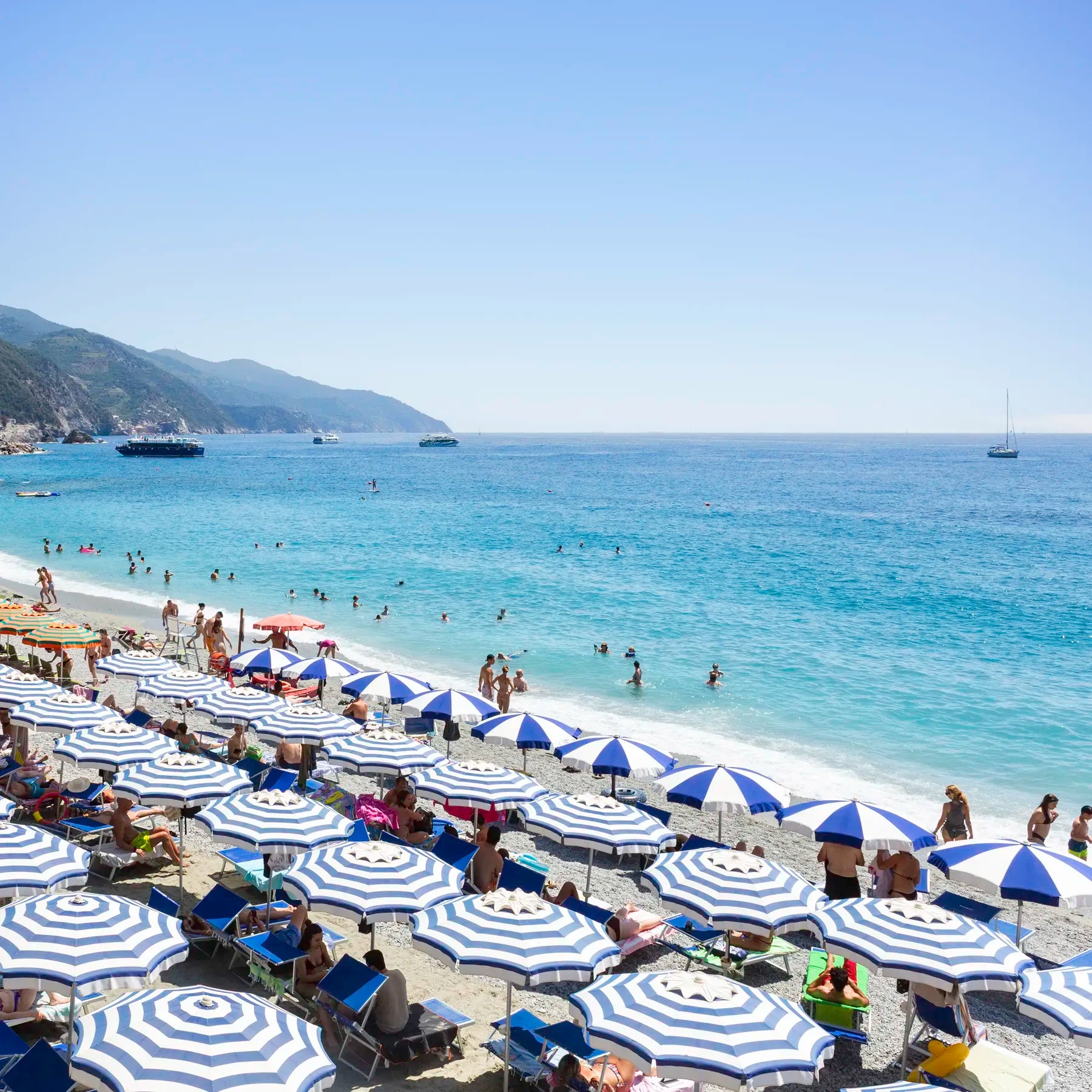 Beach scene with blue and white striped umbrellas, people, and boats on a clear day.