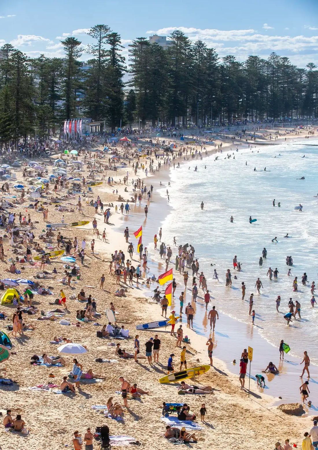 Crowded beach with people, umbrellas, and flags under a clear blue sky.