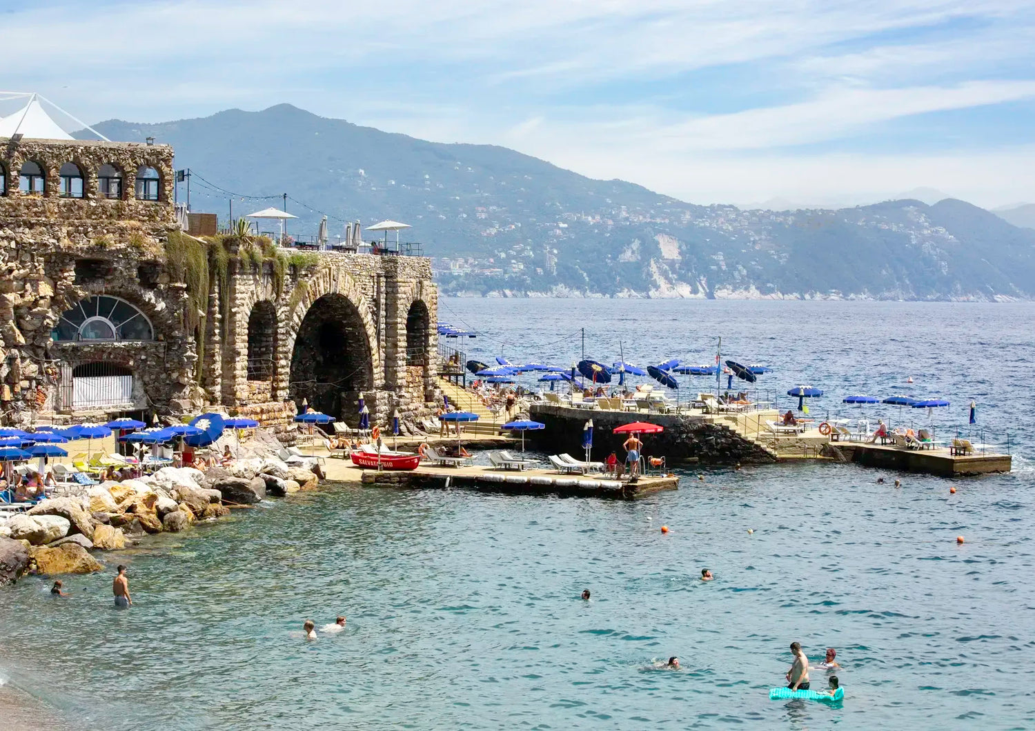 Coastal scene with stone architecture, umbrellas, and people in a body of water with mountains in the background.