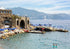 Coastal scene with stone architecture, umbrellas, and people in a body of water with mountains in the background.