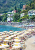 Beach scene with white umbrellas, people, and buildings on a hillside.