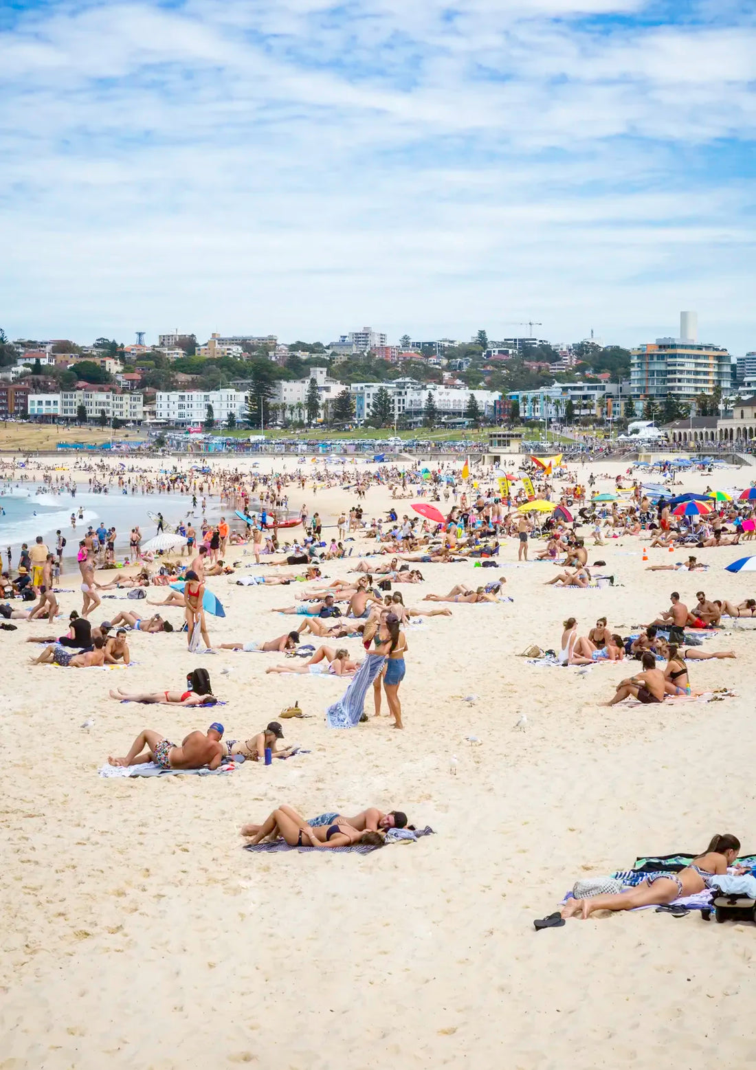 People sunbathing on a crowded beach with buildings in the background