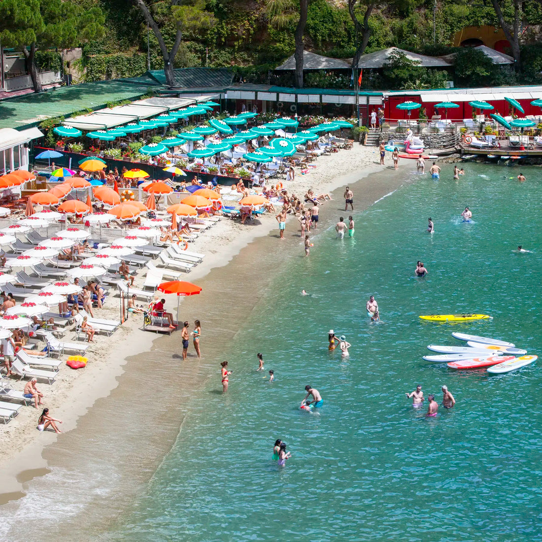 Beach scene with people swimming and sunbathing, surrounded by umbrellas and boats.