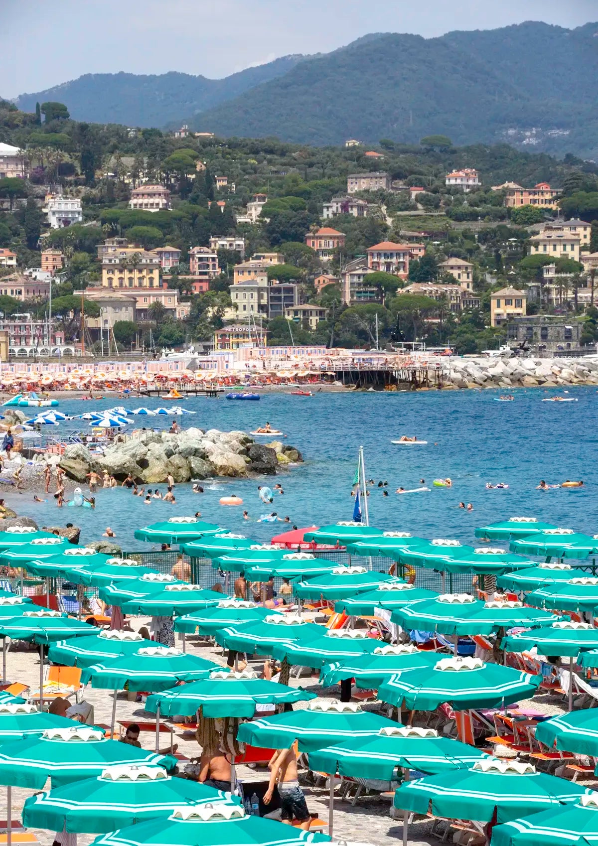 Beach scene with green umbrellas, people, and a coastal town with buildings on a hillside.