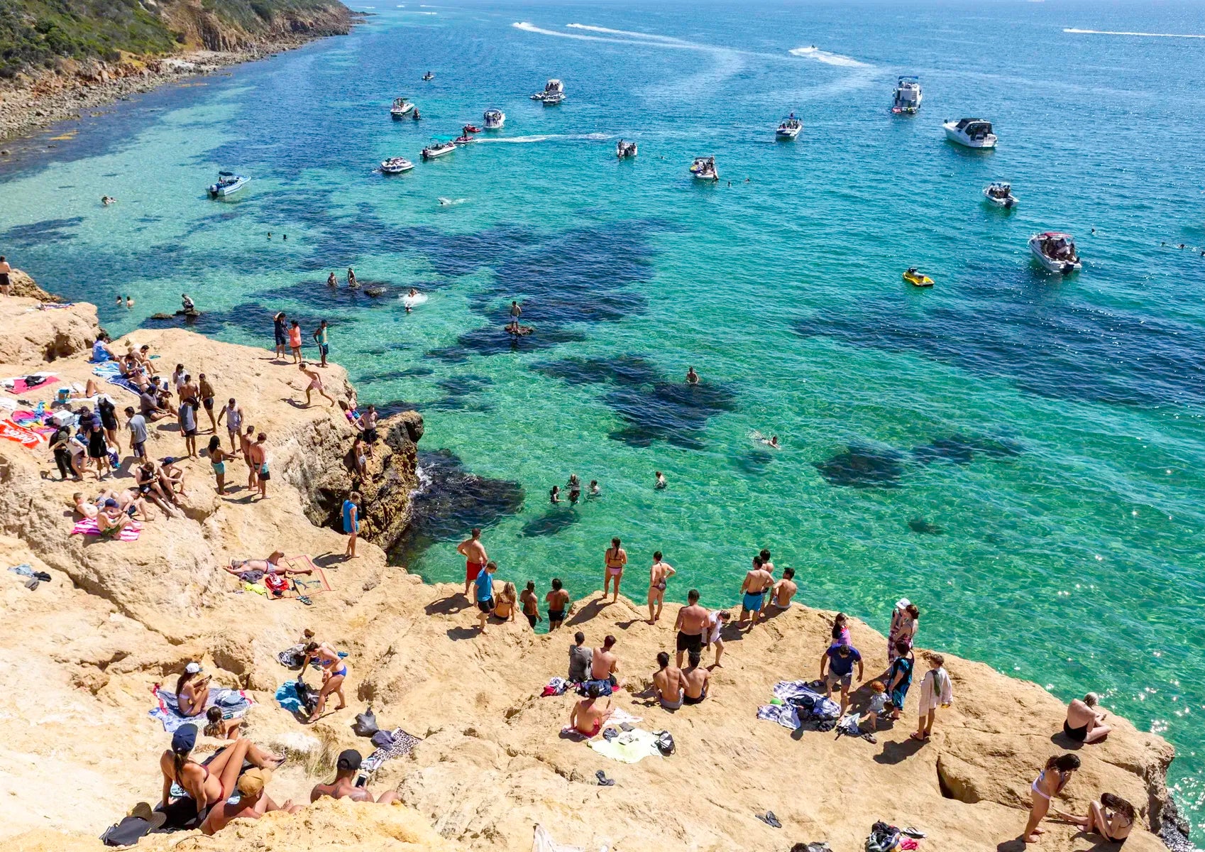 People enjoying a sunny day at a rocky beach called The Pillars in Mount Martha with clear blue water and boats.