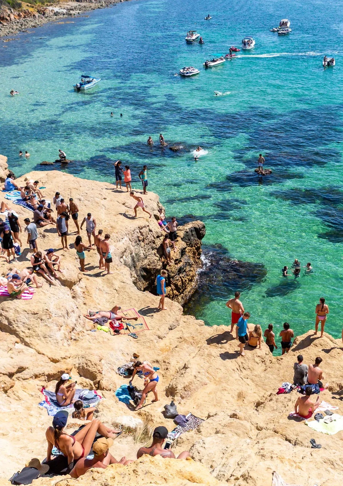 People enjoying a sunny day at a rocky beach called The Pillars Mount Martha with clear blue water and boats.