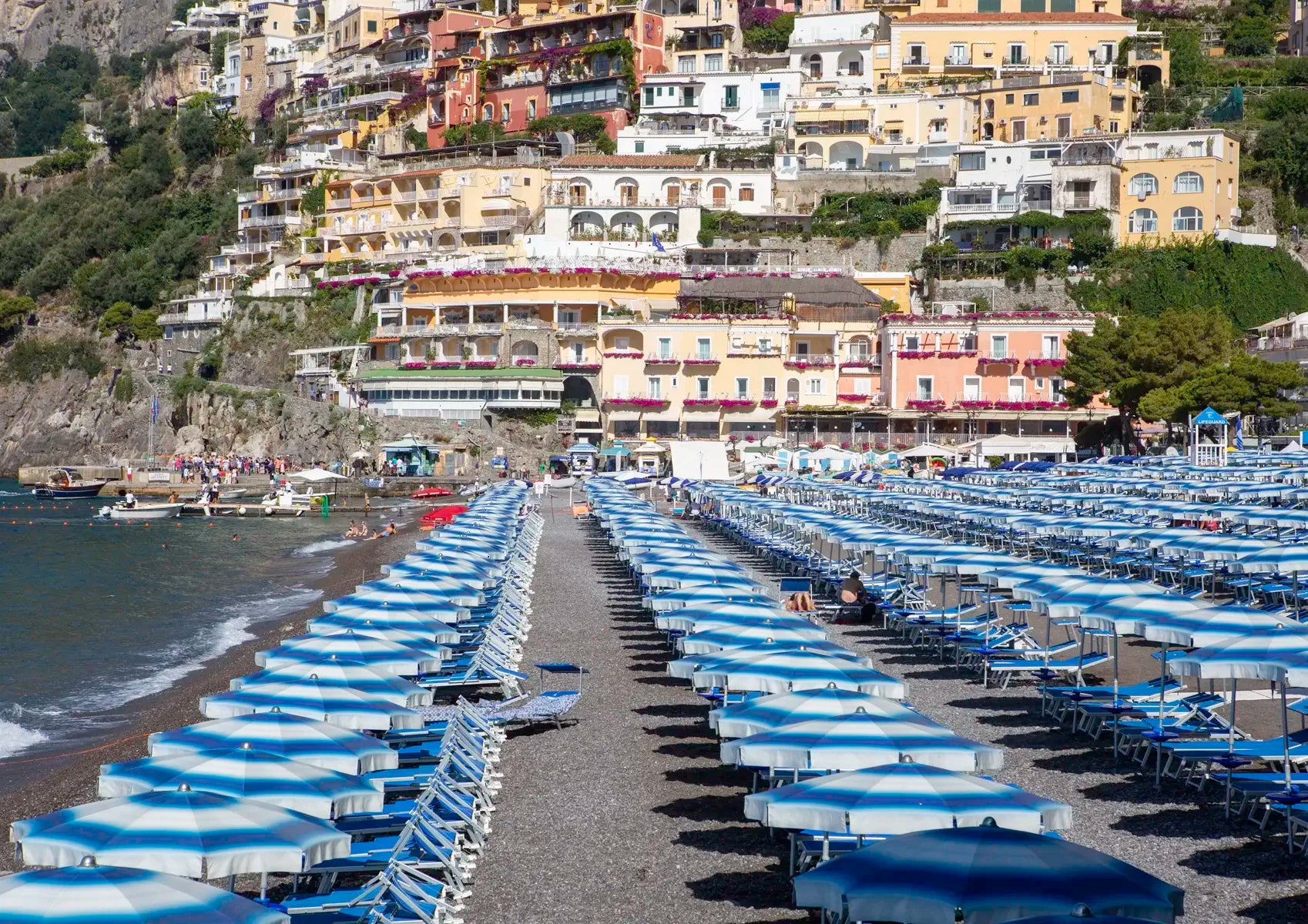 Beach with rows of blue umbrellas and chairs, buildings in the background.