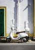 White scooter parked against a white wall with a green door.