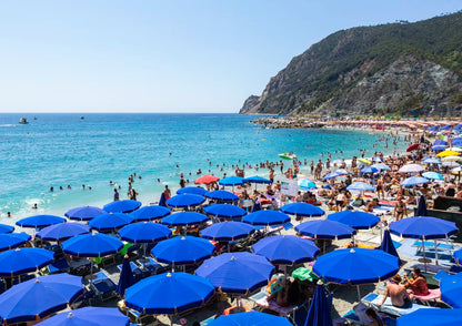 Beach scene with many blue umbrellas and people near a mountainous coastline.
